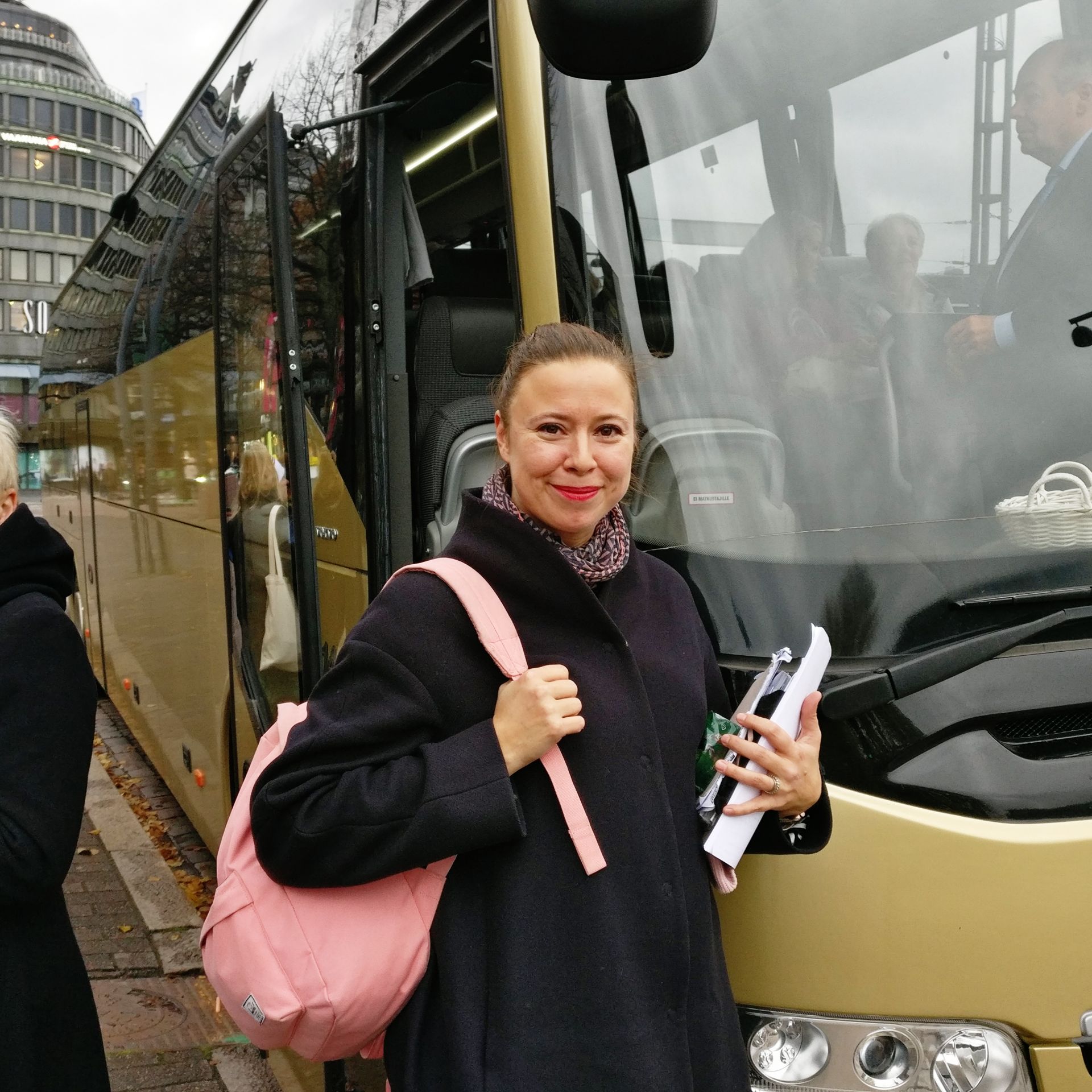 Kvinna i 30-40 års åldern med rosa ryggsäck som står framför en buss.