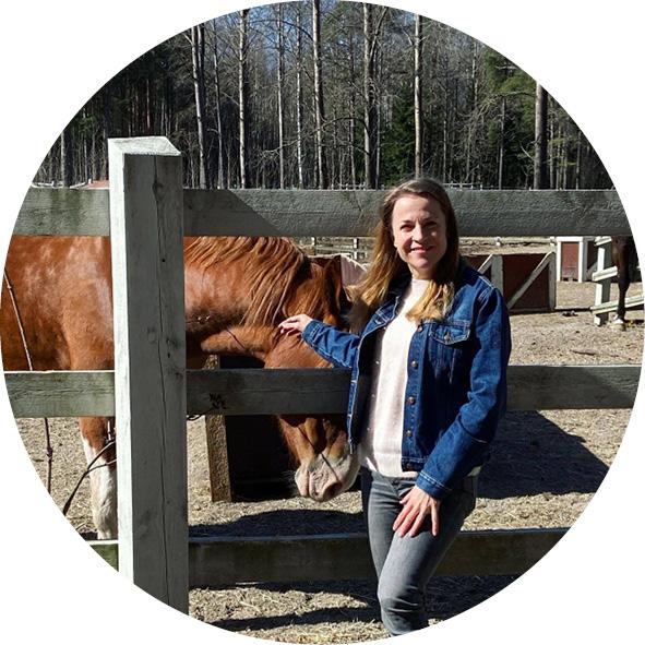 Anna i jeansjacka poserar vid ett staket och där syns en brun häst i bakgrunden.